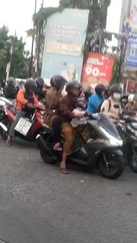 The video captures a street scene with several motorcycles and trucks. In the foreground, two people on motorcycles are stopped, with one wearing an orange jacket and the other wearing a black jacket and helmet. Behind them, more motorcycles are visible, some with riders and some empty. Further back, a white tow truck with "AUTO SECO" written on its side is parked on the right. Another truck, a white Isuzu, is seen with a person standing next to its open driver's side door. A black SUV is also present, in front of a truck with a black tarp. The environment appears to be an urban or suburban street with some greenery and buildings in the background. The overall impression is a traffic situation, possibly a slowdown or a temporary stop.