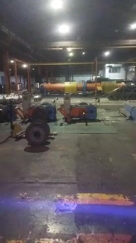The video shows an industrial setting, possibly a workshop or factory floor. The lighting is dim, with overhead lights providing illumination. In the foreground, a large tire is resting on the ground. Behind it, there are two large industrial machines, one blue and one orange, with what appear to be tires or large circular components attached to them. These machines are positioned on raised platforms. Further in the background, there is a long, multi-colored piece of machinery, predominantly orange and yellow, and several other industrial machines and equipment. There are also people visible in the background, appearing to be working around the machinery. The floor is concrete and marked with yellow and black lines. The overall environment suggests an active industrial space.