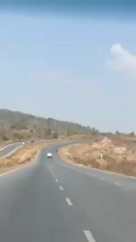 The video depicts a view from inside a moving vehicle, looking out onto a winding highway. The road is asphalt with white dotted lines marking the lanes. A white car is visible in the distance on the same side of the road, moving in the same direction.  Next to the car, a person is riding a motorcycle. The landscape to either side of the highway is arid and sparsely vegetated, with low hills dotted with trees and shrubs. The sky is light blue and hazy. The camera is positioned at the front of the vehicle, capturing the view through the windshield, and the dashboard is visible at the bottom of the frame. A piece of graffiti in pink is briefly visible at the start of the clip.