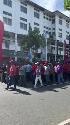 A crowd of people, many wearing red shirts and red hats, march down a city street. Red flags are visible amongst the crowd. The buildings lining the street are several stories tall and mostly white. A palm tree is visible near the front of the crowd. Some people in the crowd are talking on cell phones. Some are carrying umbrellas.