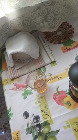 The video presents an overhead view of a table in a domestic setting, brightly illuminated by natural light entering from the left. A white, lacy fabric, possibly a curtain or part of a chair, is visible on the far left. The table is covered with a white tablecloth featuring colorful illustrations of various food items and herbs, including red and yellow bell peppers, black and green olives on a branch, rosemary, and garlic cloves. Text such as "Pepper," "Olive," "Pumpkin" (partially visible), and "Spices" is discernible on the tablecloth.

Positioned on the tablecloth are a folded, light-brown striped towel, topped with a smaller, white folded fabric. Adjacent to these, a clear glass mug sits empty. In the background, partially visible, are some dark, round objects, possibly cookies or nuts.

At the 0:01 mark, an adult hand enters the frame from the top, holding a dark-colored French press. The hand carefully positions the French press over the empty glass mug. From 0:02 to 0:06, a clear, amber-colored liquid, resembling tea or an herbal infusion, is poured steadily from the French press into the mug, filling it. The hand then lifts the French press, concluding the pouring action at 0:06, leaving the mug full. The scene depicts a simple act of preparing a beverage.