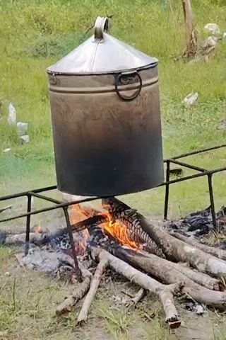 A metal pot with a lid is suspended over a wood fire in a grassy area. The pot is large and cylindrical, with a conical lid and a handle on top. It is darkened with soot from previous use. The fire is burning beneath the pot, with flames visible between the logs. The pot sits on a metal frame, holding it securely above the fire. The surrounding area is green grass, with some trees visible in the distance. There are some small pieces of litter on the ground as well. Smoke rises from the fire.