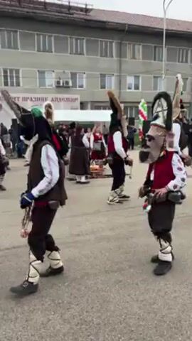 A group of people in traditional Bulgarian costumes are performing a ritual dance in front of a gray concrete building. The dancers wear elaborate masks and costumes, including bells and animal skins. They move in a synchronized manner, creating a rhythmic sound with the bells. A Bulgarian flag is visible among the dancers. In the background, a crowd of people watches the performance. A sign in Bulgarian is displayed on the building. The scene is outdoors during daylight hours.