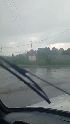 The video shows a view from inside a vehicle driving on a wet, paved road. The sky is overcast and it appears to be raining or has recently rained, as evidenced by the puddles and reflections on the road. In the distance, there is a white structure with a light-colored object on top, possibly a statue, with a utility pole to its right. The background consists of greenery, trees, and a fence line. Power lines are visible stretching across the sky. The overall atmosphere is gloomy due to the weather.