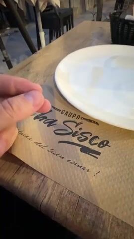 The video shows a hand making an Italian hand gesture, specifically the "pinched fingers" gesture, in front of a place setting at a restaurant. The place setting includes a paper placemat with the name "Grupo Cha SisCo" printed on it in a stylized font, with the phrase "placer del buen comer!" written below. A white plate is placed on top of the placemat. The table is wooden, and there is a black basket visible in the background. Other chairs and a table leg can be seen behind the setting, suggesting an outdoor dining area. The lighting appears to be a mix of natural and artificial. The person is likely making the gesture playfully towards the restaurant name or the concept of "good food".