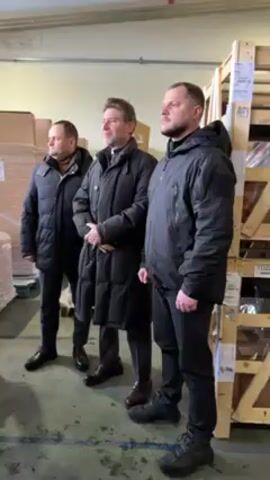 Three men in coats stand side-by-side in an industrial setting. The man on the left wears a black puffer jacket, black pants, and black shoes. The man in the middle wears a long black coat, black pants, and brown shoes. The man on the right wears a black jacket, black pants, and gray shoes. They stand in front of stacks of boxes and wooden crates. The floor is painted green and the walls are light gray brick. The lighting is bright and even.