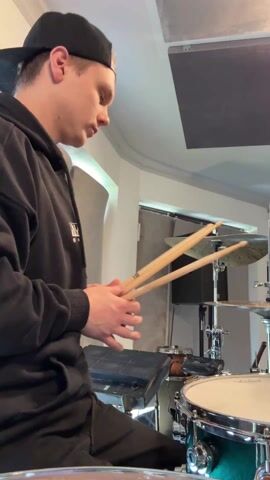 A fair-skinned, light-haired man in a black hoodie and black cap is drumming on a drum kit in a soundproofed room. The drum kit includes a snare drum, cymbals, and an electronic drum pad.  He strikes the drums with wooden drumsticks in his hands, moving them rhythmically. The room is sparsely decorated, with gray soundproofing panels on the walls. A speaker and other musical equipment are visible in the background. The lighting is bright and even.