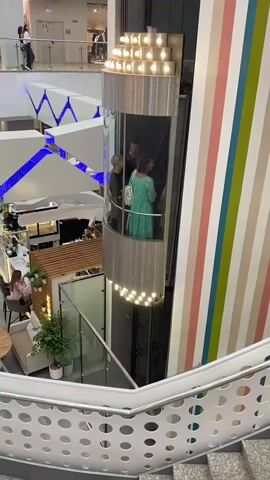 The video shows a glass elevator in a shopping mall. Inside the elevator, two women are visible. The woman on the left is wearing dark clothing and the woman on the right is wearing a green dress. The elevator is ascending. Below the elevator, a cafe is visible with several tables and chairs. A person is seated at one of the tables, facing away from the camera. The mall has modern architecture with white geometric designs on the ceiling, illuminated by blue lights. A colorful striped wall is on the right side of the frame. The view is from a higher level, looking down on the elevator and the cafe.