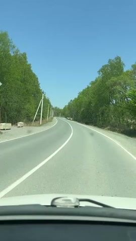 The video shows a car driving down a straight asphalt road. The road is flanked by green trees on either side, creating a kind of tunnel effect. Power poles are sparsely placed along the left side of the road. The sky is clear and blue. A vehicle is parked on the side of the road in the distance. The windshield and wipers of the car from which the video is being taken are visible in the lower part of the frame.