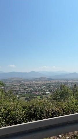 The video captures a landscape view from a high vantage point. The sky is clear blue with a few wispy clouds. In the foreground, lush green trees and foliage are visible, some with thin trunks and leafy branches reaching upwards. A white barrier or railing runs along the bottom of the frame, suggesting the viewpoint is from a road or elevated platform. In the middle ground, a town or city spreads out, with buildings and structures visible amidst more greenery. In the background, a series of rolling hills or mountains recede into the distance, their outlines softened by atmospheric haze. The overall impression is of a sunny day with a panoramic vista of a town nestled in a valley surrounded by mountains. The camera appears to be moving slowly, possibly panning or traveling along a route, as indicated by the subtle shifts in the foreground foliage and the overall perspective. There are no people or discernible actions within the frame.