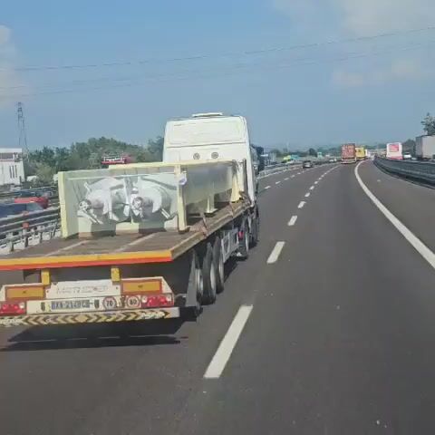 A semi-trailer truck, with a white cab, is driving on a highway. The truck is carrying a large, light-colored object secured to a flatbed trailer. The object appears to be industrial machinery, possibly with cylindrical components and metal frameworks. The trailer has yellow and black reflective markings and a license plate that reads "XA 214BC". The highway has multiple lanes, separated by white dashed lines. Other vehicles, including cars and trucks, are visible on the highway in the distance and in adjacent lanes. The sky is clear and blue with scattered clouds. The overall environment appears to be a sunny day on a busy highway.