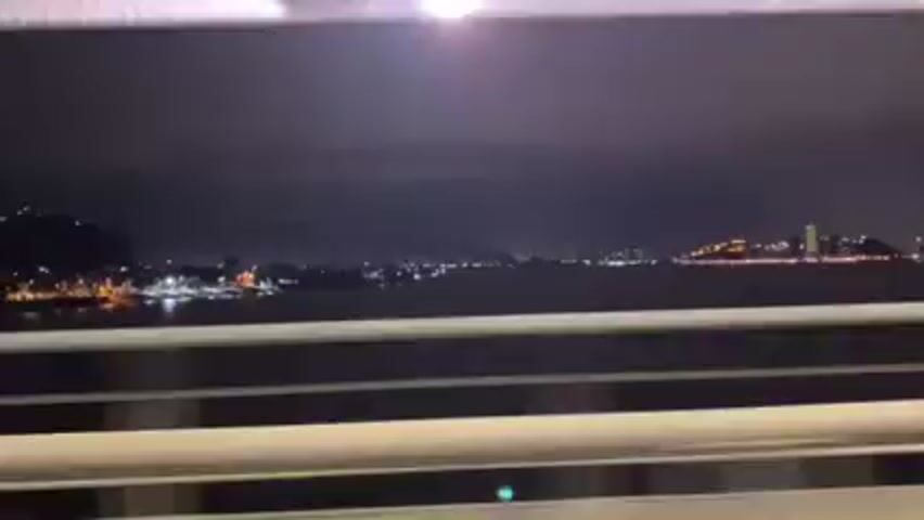The video shows a nighttime view of a city skyline across a body of water, as seen from a moving vehicle on a bridge. The skyline is illuminated with various lights, indicating buildings and infrastructure. The water reflects some of the city lights, creating a shimmering effect. The bridge's railing is visible in the foreground, blurring as the vehicle moves. The sky is dark, with some cloud cover obscuring the view of the horizon. The overall atmosphere is dark and quiet, with the city lights providing the only source of illumination.