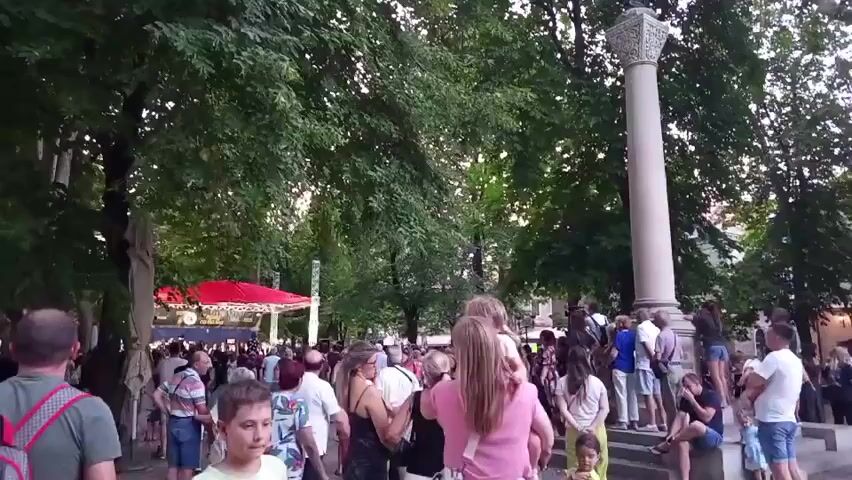The video shows a crowd of people gathered outdoors in a park-like setting, possibly for an event. In the background, a red tent or stage is visible, suggesting a performance or market. To the right, a tall, ornate column stands, topped with a statue. Lush green trees with dense foliage form a canopy over the area, creating shade. The lighting indicates it is either late afternoon or early evening. People of various ages are present, dressed in casual summer clothing. Some are standing and observing, while others appear to be moving through the crowd. The overall atmosphere seems to be lively and social, with many individuals engaged in conversation or watching the activity.