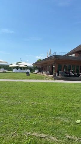 The video features an outdoor scene on a bright, sunny day. The camera pans slowly from right to left across a large, well-maintained grassy area. To the right, a modern building with an outdoor patio is visible. In the midground, several large white umbrellas provide shade over multiple white tables, which are covered with white tablecloths and surrounded by chairs, suggesting an event setup. A light-colored gravel path winds through the green lawn, branching into two directions. As the camera pans further left, more trees and greenery become visible in the background, along with a white statue or pillar positioned near the path. The sky is clear blue with scattered white clouds. No people are distinctly visible or performing specific actions.