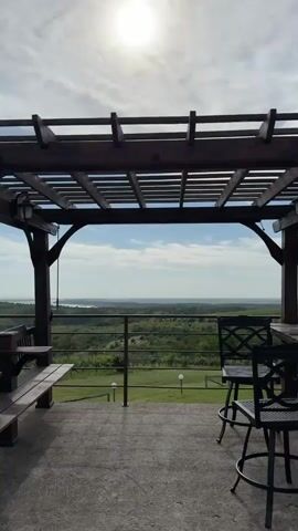 The video shows a view from under a wooden pergola structure.  The foreground features a concrete patio with a wooden bench and black metal chairs.  Beyond a metal railing, there's a gentle slope of grassy field with small light fixtures.  In the distance, a green, hilly landscape stretches out towards a wide body of water under a blue sky with some white clouds.  The camera zooms slightly to better highlight the river. The sun is visible through the pergola's slats.
