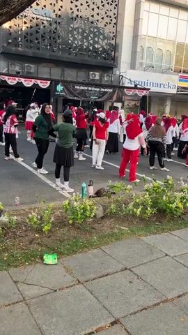 The video shows a group of people, mostly women wearing hijabs, participating in an outdoor exercise class or synchronized dance. They are dressed in athletic wear, with many wearing red and white outfits, possibly indicating a national theme or event. The participants are spread out on a paved surface, appearing to be following a leader or instructor who is not fully visible. Some are performing arm movements, while others are in a standing pose. In the background, there are buildings with storefronts, including one with a sign that appears to read "Asminareka." A planter box with greenery is in the foreground, and a sidewalk with interlocking pavers is also visible. The overall scene suggests a public event focused on fitness or community activity.
