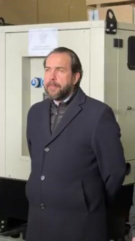 A static, eye-level indoor shot shows a man standing in front of a large generator. The man has dark hair, a beard, and is wearing a navy blue overcoat, a black zip-up vest, and a light blue collared shirt. He has his hands clasped behind his back and is looking slightly to the right of the camera.  Behind him is the generator, which is light beige in color.  It has several blue electrical outlets visible on the front panel. A white sticker is affixed at the top center of the generator, with text printed in Polish. The background appears to be a warehouse or storage area, with boxes stacked behind the generator.