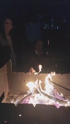 A close-up shot shows a campfire at night, with two figures visible in the dark background. The fire is burning brightly, with flames and sparks rising. The figures are out of focus, but one appears to be standing and the other to be seated or leaning. The fire is contained within a metal fire pit.