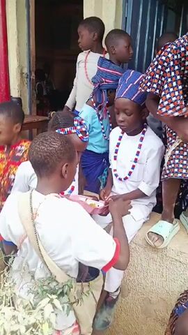 A group of children are gathered outside a building, eating from small plates. Several are wearing traditional clothing, including white shirts and pants, beaded necklaces, and headbands or hats with blue and red patterns. One child has a bag filled with what appears to be leafy green plants. They are sitting on or near a low wall or ledge, and more people are visible inside the building. The environment suggests a community gathering or celebration.