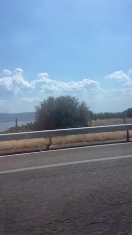 A static shot looking out from a moving vehicle. A highway guardrail runs diagonally across the frame from left to right. Beyond the guardrail is a body of water, likely a lake, with a distant shoreline dotted with trees. The sky above is a clear blue with fluffy white clouds.