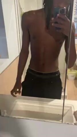 A slender, shirtless person with dark skin and dreadlocks is taking a mirror selfie in a bathroom. They are wearing dark pants with a visible black and green waistband. The bathroom has beige tile walls and a white sink and counter. The person blows out a plume of smoke. The phone case is blue and purple. A shower door is visible behind the person.