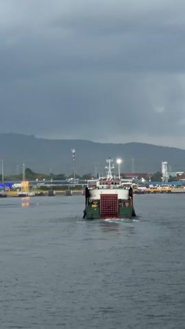 A ferry boat is sailing away from the camera on a body of water. The sky is cloudy and dark, suggesting it may be overcast or close to evening. In the background, there are hills or mountains, and along the shoreline, there are buildings and structures, including what appear to be industrial facilities and possibly a port or harbor. Another larger ship is docked on the right side of the frame. The ferry boat itself is green and white, with a red cargo container visible on its deck. The water is calm with ripples from the boat's movement.