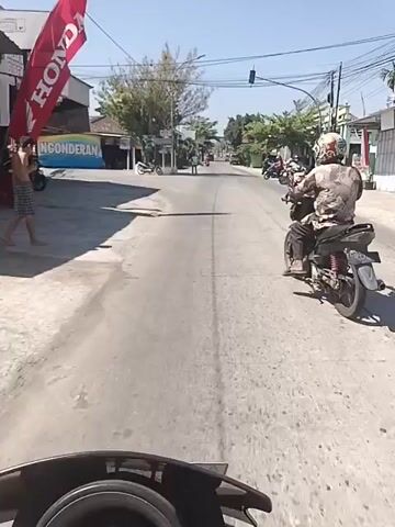 The video shows a first-person perspective of someone riding a motorcycle down a paved road on a sunny day. The rider is following another motorcycle with a person wearing a patterned shirt and helmet. The environment appears to be a town or village, with buildings lining the road on both sides. Signs are visible on the buildings, including one that says "NGONDERAN." A large archway with writing on it is seen in the distance, and a cell tower is behind it. Other motorcycles and a car are also visible on the road. The scene is dynamic, with the rider's movement creating a sense of forward momentum.