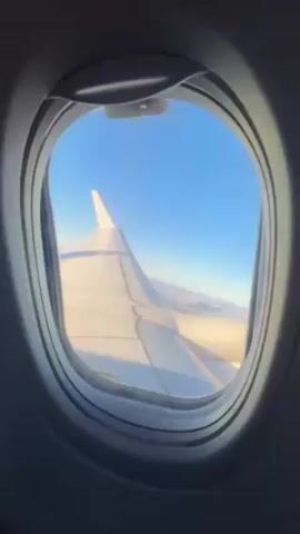 The video shows a view out of an airplane window. The camera is static, looking out at a clear blue sky with the plane's wing visible in the frame. The wing is white and appears to be in flight. The airplane window is oval-shaped and has a latch at the top. The interior of the plane is dark compared to the bright exterior view.