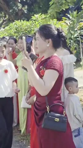 The video shows a group of people, primarily women, some dressed in red traditional clothing. One woman is wearing a yellow face mask. They appear to be part of an event or celebration. Some people are holding phones and filming or taking photos. A man wearing a white shirt speaks to the camera. A white van with the words "SANG T" is visible. The background includes green foliage and some buildings.