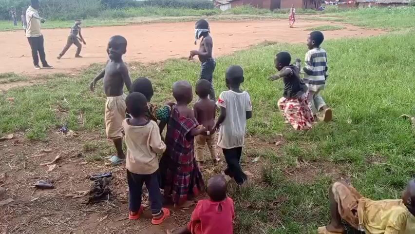A group of about a dozen children are playing in a grassy area next to a dirt field. One child has a blindfold on. Another child is running across the field. An adult is standing on the edge of the field watching. There are a few simple houses visible in the background. The children are laughing and running around. In the distant background, a mountain can be seen.
