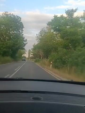 The video shows a view from inside a vehicle as it travels on a paved road. On the left side of the road, there are trees and vegetation, with a white line marking the lane. On the right side, there is more vegetation, including trees and tall grass, and a white post with reflective markers. The road curves to the left and ahead, a dark-colored car is visible in the distance. The sky is cloudy and appears to be either early morning or late afternoon. In the distance, a tall, thin tree or structure is visible.