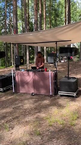 The video shows a DJ set up in a wooded area, likely in Mednyi Zavod, Russia. A man, dressed in a leopard print shirt, stands behind a DJ booth covered by a large canopy. He is actively engaged in mixing music, as indicated by his posture and the equipment in front of him, which includes turntables and a laptop. The booth itself is draped with a brown fabric and adorned with LED light strips, adding a festive touch to the outdoor setting.

Surrounding the DJ booth are several large speakers, suggesting a public event or party. The scene is set amongst tall pine trees, with dappled sunlight filtering through the branches, indicating it is daytime. The ground is covered in pine needles and dry leaves, typical of a forest floor. In the background, more trees and a clearing can be seen, hinting at a larger gathering or festival area. The overall atmosphere appears relaxed and celebratory, with the natural beauty of the forest providing a unique backdrop for the musical performance. The weather seems pleasant, conducive to an outdoor event. No visible text is present in the video.