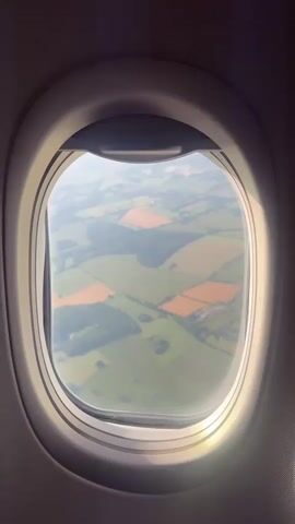 The video shows a view through an airplane window of a patchwork of green and brown agricultural fields below, possibly farmland. The window frame is visible, along with the interior of the aircraft cabin. The lighting suggests daytime. The video is short, with no discernible actions or interactions taking place.