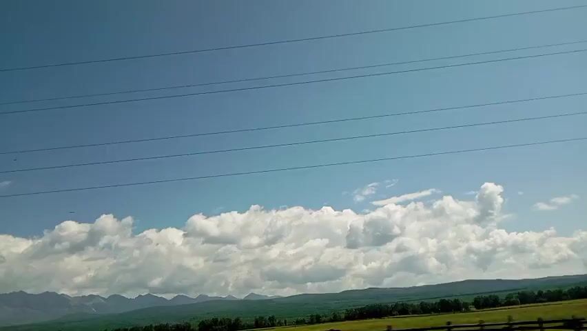 The video shows a wide shot of a rural landscape on a clear day. The sky is a bright blue with several power lines stretching horizontally across the frame. Below the sky, fluffy white clouds form a dense line above a distant mountain range. The mountains are green and appear to be covered in trees. In the foreground, there is a field of green grass, and a fence is visible on the right side of the frame. A power pole is also visible on the right side of the frame, with wires extending from it. The camera appears to be moving slowly, likely from a vehicle.
