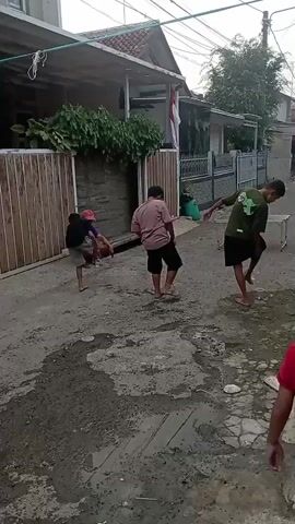 The video shows a group of children playing in a narrow, unpaved alleyway between houses. The ground is uneven and appears to be a mix of dirt and some sort of dark gravel or aggregate, with several puddles of standing water.

In the foreground, a child wearing a red shirt and black shorts is facing away from the camera and moving to the left.

Further back in the alley, there are four individuals. On the left, a person wearing a dark shirt and a red cap appears to be holding a brown object, possibly an animal or a bundle, and is leaning over. Next to them, a child in a light pink shirt and black shorts is bent over, with their left leg raised. To their right, a child in a green t-shirt and black shorts is also bent over, with their right leg extended backwards. Further to the right, a person wearing a blue headscarf and a beige outfit is standing and looking towards the child in the red cap.

The buildings lining the alley appear to be simple, multi-story houses with tiled roofs and some vegetation. There are fences and walls separating the properties. The overall impression is a casual, outdoor activity in a residential area.