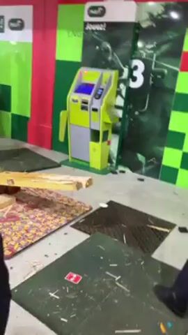 The video shows the aftermath of what appears to be vandalism or destruction within a retail space. A person in dark clothing bends over amid scattered debris and damaged flooring. A bright green and grey kiosk or machine stands relatively undamaged. The walls are painted in a pattern of green and red with the word "Jouez!" visible. In the background, a structure of metal bars is apparent, leaning against the wall. The floor is covered with pieces of carpet, metal grates, wood, and other scattered materials.
