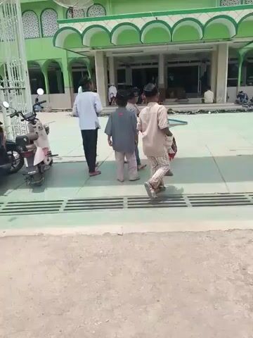 The video shows several children standing outside a mosque. Three boys stand near a set of metal grates in the foreground. One boy wears a white shirt and dark pants, one boy wears a brown shirt and dark pants, and one boy wears a grey shirt and tan pants. All the boys wear a black hat, and they are all barefoot. The boy in the white shirt appears to be giving the other boys money. To the left of the boys is a moped. In the background, more children and adults are walking into the mosque. Many pairs of shoes are lined up in front of the entrance. The mosque is painted light green, and the pillars are white.