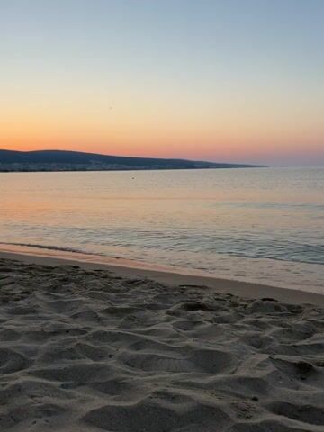 This is a video of a beach at sunset. The sand in the foreground is a light brown color and has ripples and textures from the wind. The ocean is calm, with small waves gently rolling onto the shore. The water is a soft, muted pinkish-orange color, reflecting the sunset. In the distance, a dark, hilly coastline is visible. The sky is a gradient of colors, transitioning from a pale blue at the top to a warm orange and pink near the horizon. There are no people or objects visible in the video. The video is handheld and has a slight sway.