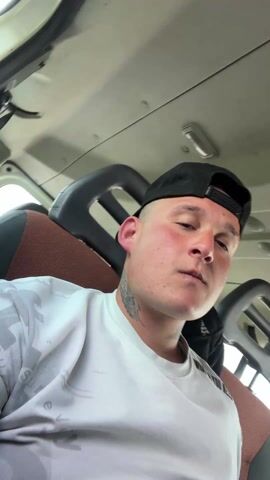 The video shows a fair-skinned man with a buzz cut and a black baseball cap sitting in the passenger seat of a vehicle. He is wearing a white long-sleeved shirt with a gray pattern. A tattoo is visible on his neck. The interior of the vehicle is visible, with a brown seat and a gray ceiling. The camera angle shifts upward, briefly showing the vehicle's ceiling.