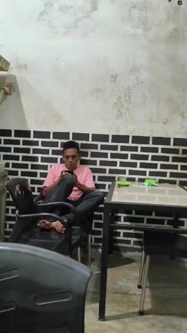 A man sits cross-legged in a black plastic chair, facing the camera and looking down at a black mobile phone he holds in his hands, seemingly playing a game. He wears a light pink t-shirt, dark jeans, and is barefoot. The chair is positioned against a wall with black and white brick patterned wallpaper. To his right, there is a table with a stainless-steel frame and a shiny gray top, on which there's a green plate. Behind the table, there is another chair. The wall above the brick pattern is off-white. A section of another black chair is visible in the lower left corner of the frame. The lighting is dim.
