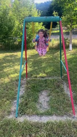 A young girl with dark hair, wearing a pink dress with a unicorn print and a colorful striped jacket, is swinging on a yellow swing set. She is wearing pink shoes and pink socks. The swing set has green and red poles and a green crossbar. The ground beneath the swing is covered in grass, with some white lines marking a path. The background is a yard with trees and green foliage. The girl is smiling and looking at the camera as she swings back and forth.