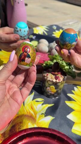 Three hands hold Easter eggs over a table with a sunflower tablecloth. One egg has an image of Jesus, one is blue with a flower pattern, and one is blue with clouds and heart imagery. The table contains food, including Easter cupcakes, eggs, greens, and other unidentified items. There are two people present, one wearing a pink sweater.