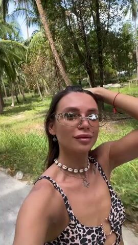 A woman is riding a scooter on a paved path in a tropical environment. She is wearing a leopard print bikini top, a white skirt, sunglasses, and a shell necklace. Palm trees and greenery are visible in the background. She is looking at the camera and adjusting her hair.