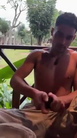 A shirtless man with a mustache sits in a chair, outside on a porch or balcony, and opens a small, dark container. He is wearing khaki-colored pants and a dark pendant on a necklace. The man has a tattoo on his left shoulder with Chinese characters. The balcony overlooks a grassy area with trees in the background. The view suggests a tropical or semi-tropical environment.  A wooden railing surrounds the balcony. A song plays in the background.