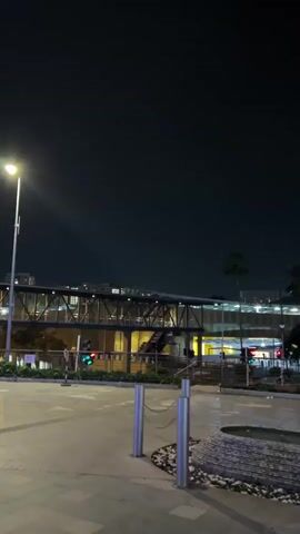 The video shows a street scene at night, illuminated by streetlights. A pedestrian bridge is visible, with palm trees lining the street. The pavement is wet, possibly from rain. There is a small circular planter in the foreground. A motorcycle drives by on the street.