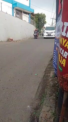 The video shows a street scene. In the foreground, a white van is driving down the road. To the right, a red sign with yellow lettering is visible. In the middle ground, two people on a motorcycle are riding away from the camera. In the background, a building with blue trim is visible, along with trees and other structures. The road appears to be paved and is in a slightly worn condition. The weather appears to be overcast or the lighting is subdued.