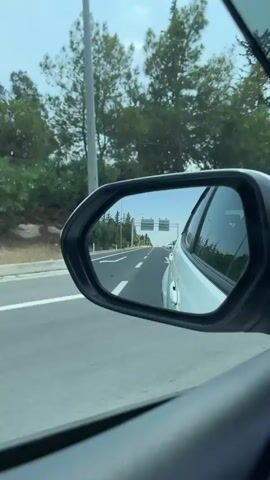 The video shows a view from inside a car, looking out the side window and into the side-view mirror. The car is in motion on a highway. The side-view mirror reflects a road stretching ahead, with trees and vegetation on either side. Another white car is visible in the reflection, slightly behind and to the right of the car from which the video is taken. The sky is a clear blue. The footage appears to be shot on a sunny day.