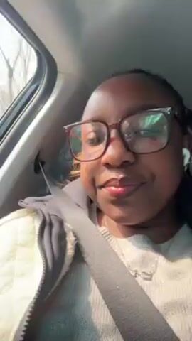 A woman sits in the passenger seat of a car. She wears glasses, a gray sweater, and a light yellow vest. She is wearing a seatbelt. She has dark hair pulled back, and a pendant necklace. The car is moving, and the video is shaky. The woman says, "Hey guys, let's go together for our sip and chat."