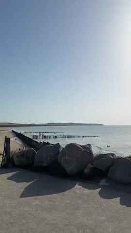 The media shows a serene beach scene in Wissant, France. The foreground features several large, gray boulders, casting long shadows on a paved walkway. Behind the rocks, a sandy beach stretches out, lined with wooden posts that form a breakwater. People are scattered along the beach and in the shallow water, enjoying the day.

The sea is calm and reflects the clear, bright sky, indicating fair weather. The sun is high, suggesting it is midday or early afternoon. In the distance, rolling sand dunes and a distant headland with a lighthouse can be seen, confirming the location as Wissant, a coastal town in northern France known for its beaches and kite-surfing.

The overall mood is peaceful and relaxed. The gentle waves, the presence of people enjoying leisure activities, and the expansive, clear sky contribute to a sense of tranquility. There is no visible text in the media.