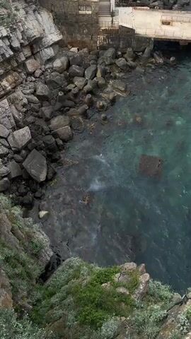 A high-angle shot shows a rocky coastline meeting clear turquoise water. Large gray rocks and cliffs surround the water's edge, some covered in green vegetation. A small concrete structure with stairs is visible in the background. The water is partially clear, revealing rocks and potential sea life below. The wind seems to be present.